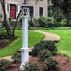 a white lamp post in front of a house on a lawn with bushes and trees