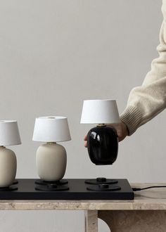 three white and black lamps on a table with one being held over by a person's hand