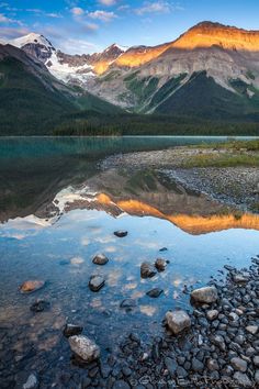 the mountain range is reflected in the still water of this lake, with rocks and pebbles on the shore