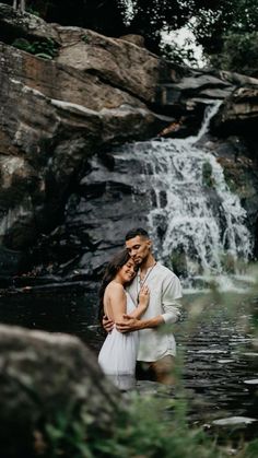 a man and woman are standing in the water near a waterfall with their arms around each other