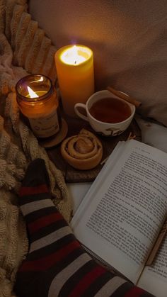 an open book sitting on top of a bed next to a cup of tea and a candle