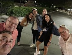 a group of people posing for a photo on the sidewalk at night time with their arms around each other