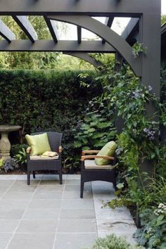 two chairs sitting under an arbor in the middle of a patio with greenery on either side