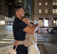 a man and woman hugging on the street at night with city lights in the background