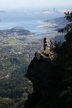 two people sitting on the edge of a cliff overlooking a city and water below them