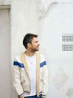 a man standing in front of a white wall wearing a tan jacket and blue jeans