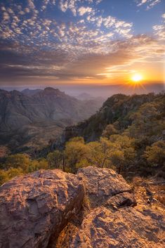 the sun is setting on top of a mountain with trees in the foreground and clouds in the background