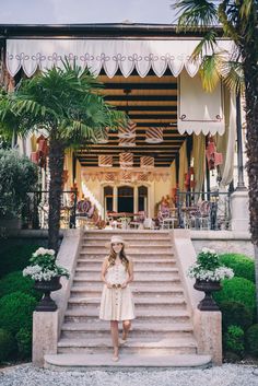 a woman standing on the steps in front of a building with palm trees and potted plants