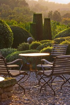 two chairs and a table sitting on top of a gravel covered ground next to bushes