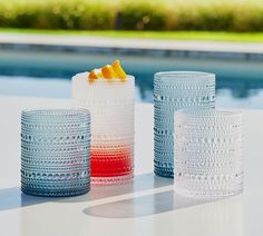 three different colored glasses sitting next to each other on a table near a swimming pool