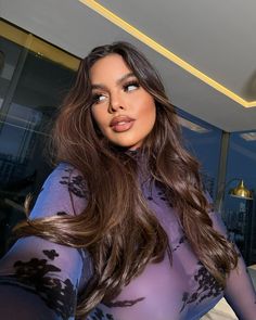 a woman with long brown hair wearing a sheer purple bodysuit and posing for the camera