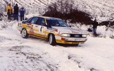 a yellow and white car driving down a snow covered road with people standing around it