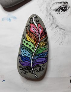 a rock with a colorful feather painted on it next to a pen and ink drawing