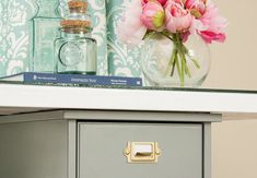 pink flowers are in a glass vase on top of a gray dresser with blue wallpaper