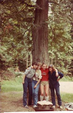 four people standing in front of a large tree with their arms around each other's shoulders