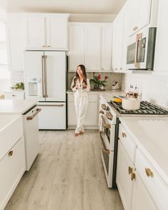 a woman standing in the middle of a kitchen with white cabinets and wood flooring