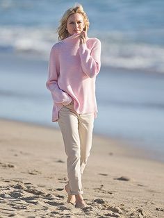 a woman is walking on the beach talking on her cell phone while wearing a pink sweater and khaki pants