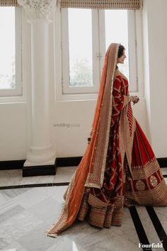 a woman in a red and gold bridal gown standing by a window with her hands on her hips