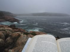 an open book sitting on top of a rocky cliff next to the ocean with waves coming in