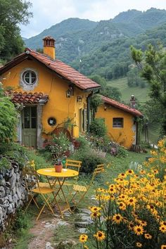 a small yellow house sitting in the middle of a lush green field with flowers around it