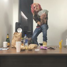 a man with pink hair sitting on top of a wooden table next to bottles and other items