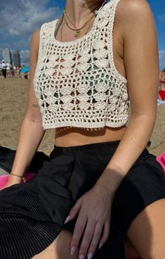 a woman sitting on the beach wearing a white crochet top and black skirt