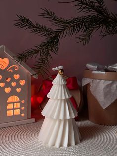 a small white christmas tree sitting next to a lit up house and gift boxes on a table