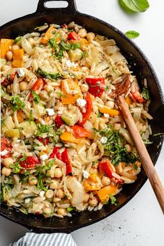 a skillet filled with pasta and vegetables on top of a white table next to a wooden spoon