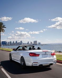 a white convertible car driving down the road next to water and cityscape in the background