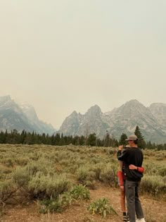 a man and woman standing in the middle of a field with mountains in the background