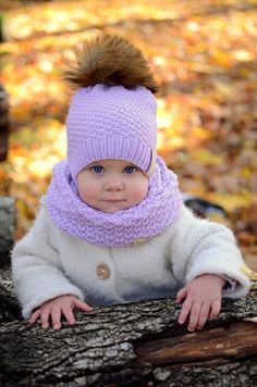 a baby wearing a purple knitted hat and scarf sitting on a tree branch in the woods