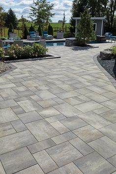 a large brick patio next to a swimming pool with lounge chairs and trees in the background