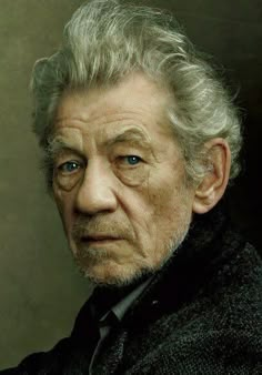 an old man with grey hair and blue eyes looks at the camera while wearing a black coat