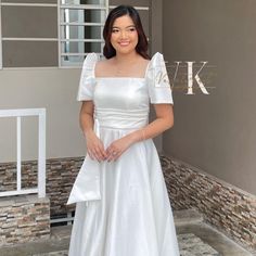 a woman wearing a white dress standing in front of a wall and posing for the camera