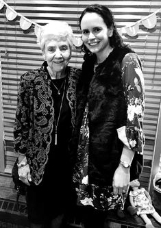 two women standing next to each other in front of a window with the blinds closed