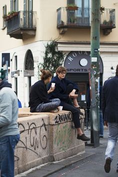 two people sitting on a wall looking at their cell phones while others walk down the street