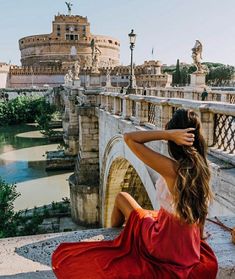 a woman in a red dress is sitting on the edge of a bridge looking at an old castle