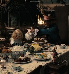 two dolls are sitting at a table with food on it and one doll is reaching for the cake