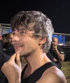 a man in black tank top standing next to other people at an outdoor event with one hand on his chin