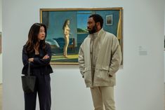 two people standing in an art gallery looking at paintings on the wall and one person holding a handbag