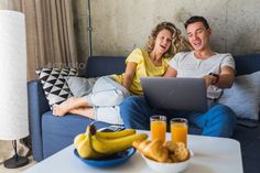 a man and woman sitting on a couch looking at a laptop computer with orange juice in front of them