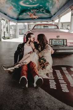 two women sitting on the ground in front of a pink car and one is kissing