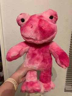 a pink stuffed animal being held up by someone's hand in front of a white wall