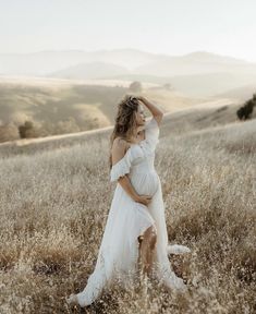 a pregnant woman in a white dress standing in a field with her hands on her head