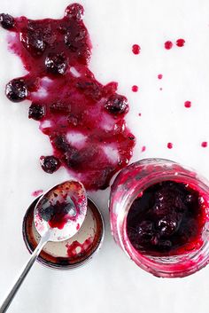 a spoon with some blueberry sauce on it next to a jar of jam and another bowl filled with berries