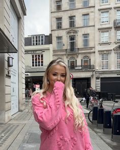 a woman with long blonde hair is standing on the sidewalk in front of some buildings