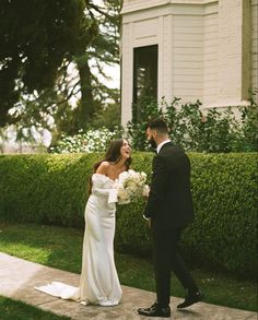 a bride and groom are walking together in front of a house with hedges on either side