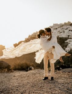 a man carrying a woman in his arms on top of a rocky hill with buildings in the background