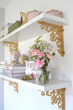 flowers in a vase on a shelf with books and other items behind it that are gold