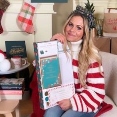 a woman sitting on a couch holding up a box with christmas decorations in the background